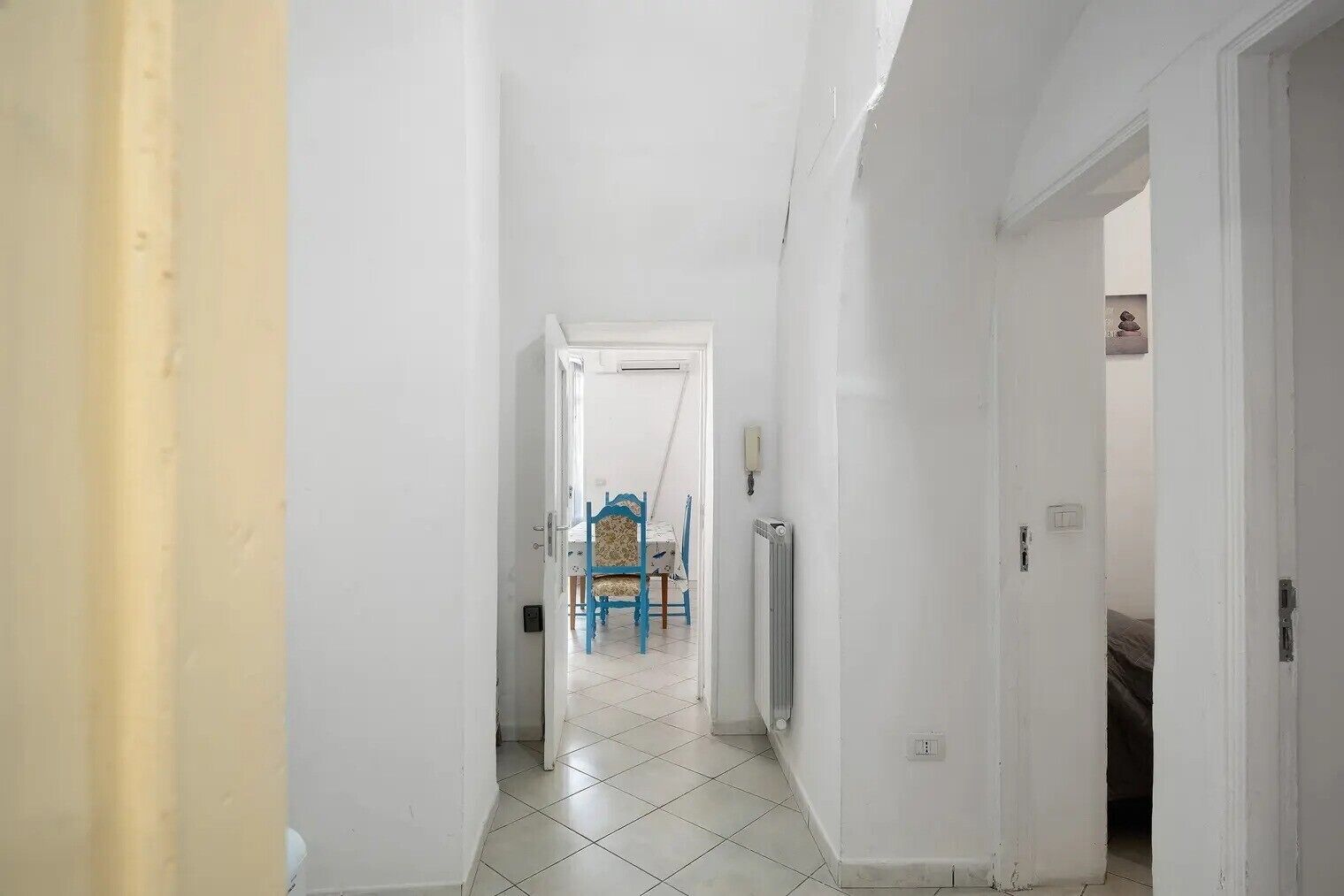 Un estrecho pasillo con paredes blancas y suelo de baldosas que conduce a una habitación con una cama y una silla.