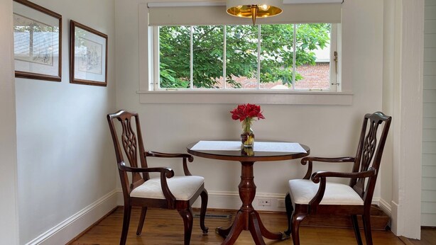 A dining area with a round table, two chairs, a vase with flowers, and a window with a view of greenery.