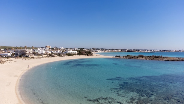 Une plage côtière aux eaux limpides, avec un rivage sablonneux et des bâtiments visibles à l’arrière-plan.