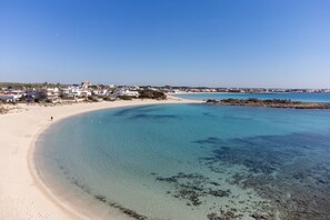 A coastal beach with clear waters, sandy shore, and buildings in the background.