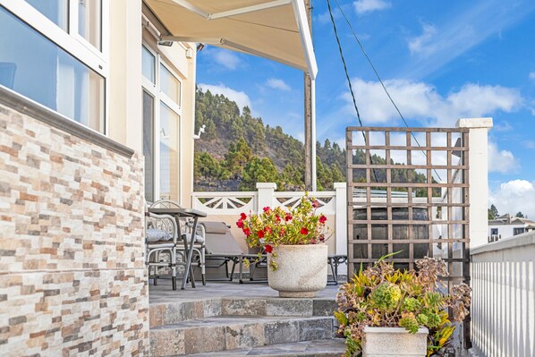 Un espace extérieur abrité, aménagé avec une table et des chaises, un mur en pierre, un pot de fleurs aux fleurs rouges et une vue sur un paysage montagneux.