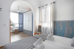 Une chambre avec deux lits, une armoire, une chaise et une vue sur l’extérieur, visible à travers une porte en arc de cercle.