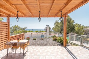 Une terrasse extérieure couverte, avec une table et des chaises, offrant une vue sur la mer et donnant sur un jardin.