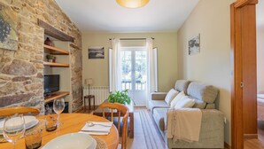 Habitació acollidora amb paret de pedra, taula de dinar amb cadires, sofà i finestra amb cortines.