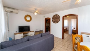 A modern living room with a grey sofa, a flat-screen TV, a ceiling fan, and a kitchen visible through an open doorway. - 2 bedroom beach front apartment in Rtina (Rtina)