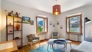 A modern living room with a wooden floor, a blue rug, a round table, two chairs, a bookshelf, and large windows with a view of mountains. - Apartment 'Casa Anna' with Mountain View and Private Terrace (Santa Maria Maggiore)