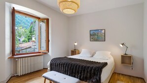 A bedroom with a bed, bench, bedside tables, a hanging light, a picture on the wall, and a window with a view of greenery and buildings. - Apartment 'Casa Anna' with Mountain View and Private Terrace (Santa Maria Maggiore)