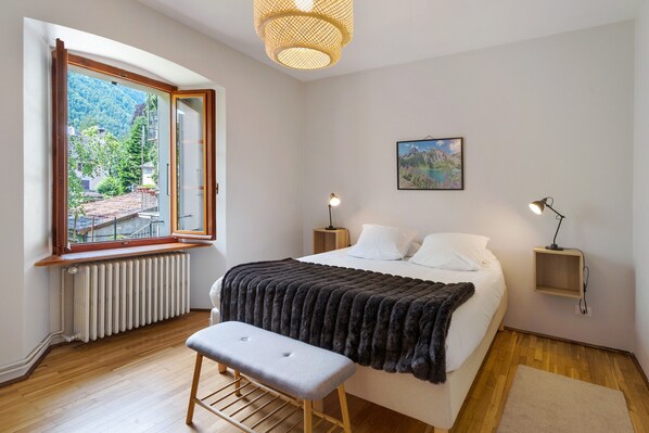 Quarto com cama, banqueta, mesas de cabeceira, luminária pendente, quadro na parede e janela com vista para área verde e prédios.