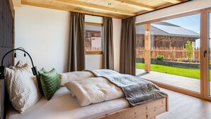 A bedroom with a wooden ceiling, large windows, and a view of a wooden fence and greenery outside.