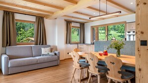 A modern kitchen with wooden beams, a dining area with a wooden table and chairs, and a grey sofa with a view of greenery outside.