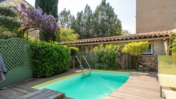 Una terraza de madera con piscina, rodeada de vegetación y un edificio de techo a tejas.