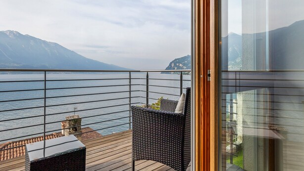 Un balcon avec vue sur un lac et des montagnes, une chaise en osier et une petite table avec un coussin.