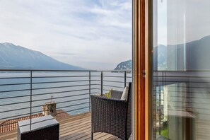 Un balcón con vista a un lago y montañas, una silla de mimbre y una mesita baja con un cojín.