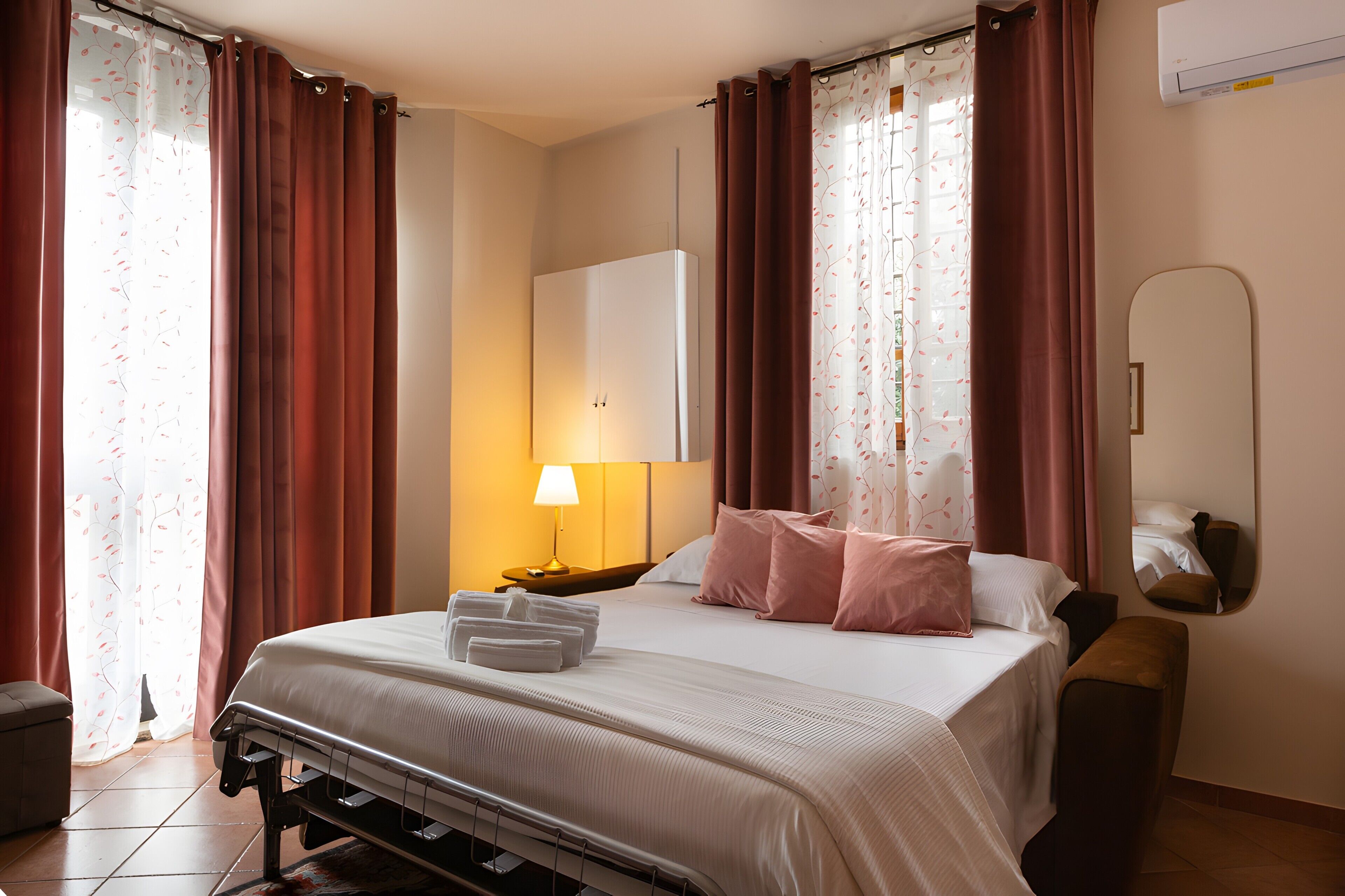 A hotel room with a bed, pillows, a bedside table, a lamp, a mirror, and a window with curtains.