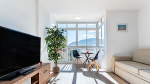 A modern living room with a flat-screen TV, a sofa, a dining table with chairs, a potted plant, and large windows offering a view of mountains. - Casa Emilio Apartment with Wi-Fi and Air Conditioning (Benalmádena)