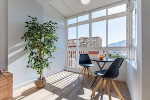 Ein Zimmer mit großem Fenster, einem Blumentopf, zwei Stühlen, einem kleinen Tisch und einem Holzkasten.