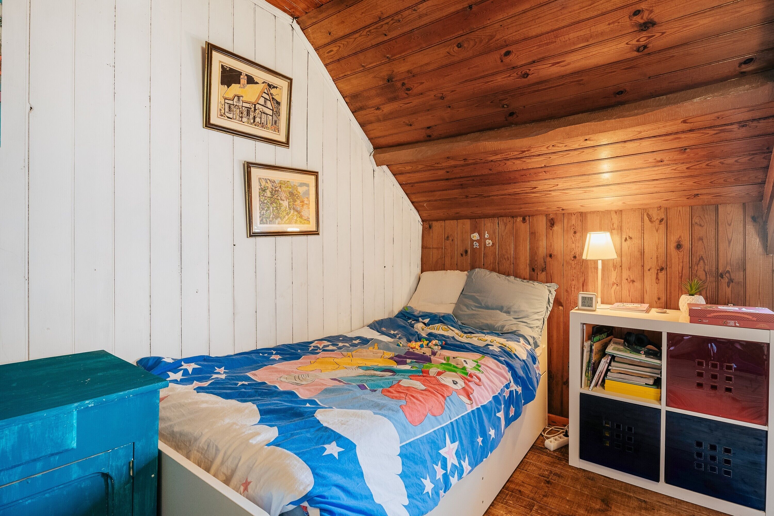 A bedroom with a wooden ceiling, a bed with a colorful quilt, a nightstand with a lamp, and a built-in bookshelf.