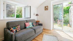 A grey sofa with colorful pillows, a lamp, and a wicker chair in a room with large windows and a sliding glass door to a garden. - Holiday Home 'Maison Pleneuf Val André Proche Mer' with Private Terrace, Private Garden and Wi-Fi (Pléneuf-Val-André)