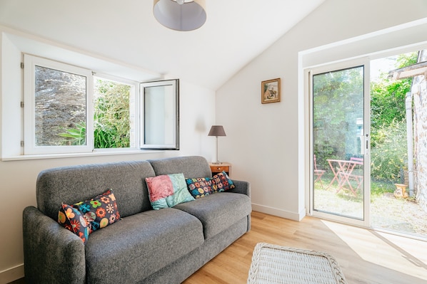 A grey sofa with colorful pillows, a lamp, and a wicker chair in a room with large windows and a sliding glass door to a garden.