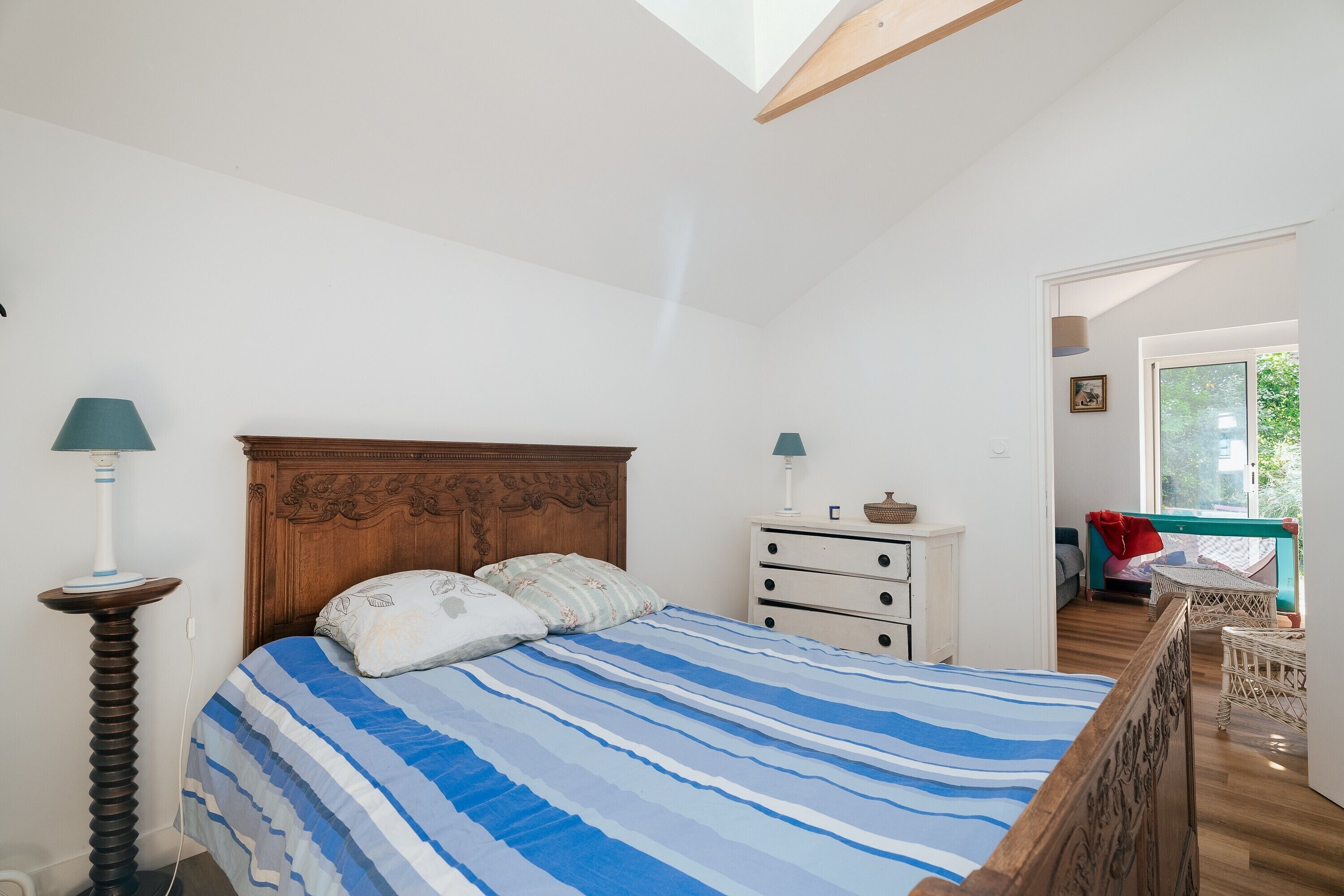 Un dormitorio con cabecera de madera, una cama con ropa de cama a rayas, una mesita de noche con lámpara y vistas a un espacio exterior.