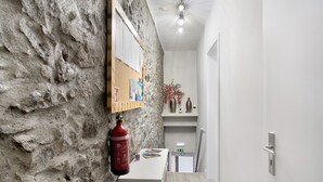 Una habitación con pared de piedra texturizada, un tablón de corcho con notas, un extintor, una mesita con un jarrón, y una puerta con manija.