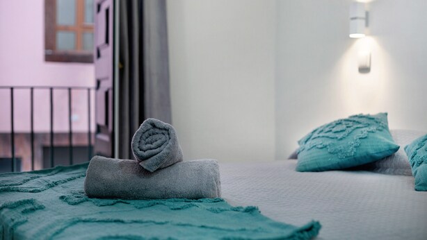 Habitación de hotel con cama, toalla enrollada, almohada color turquesa y lámpara fijada a la pared.