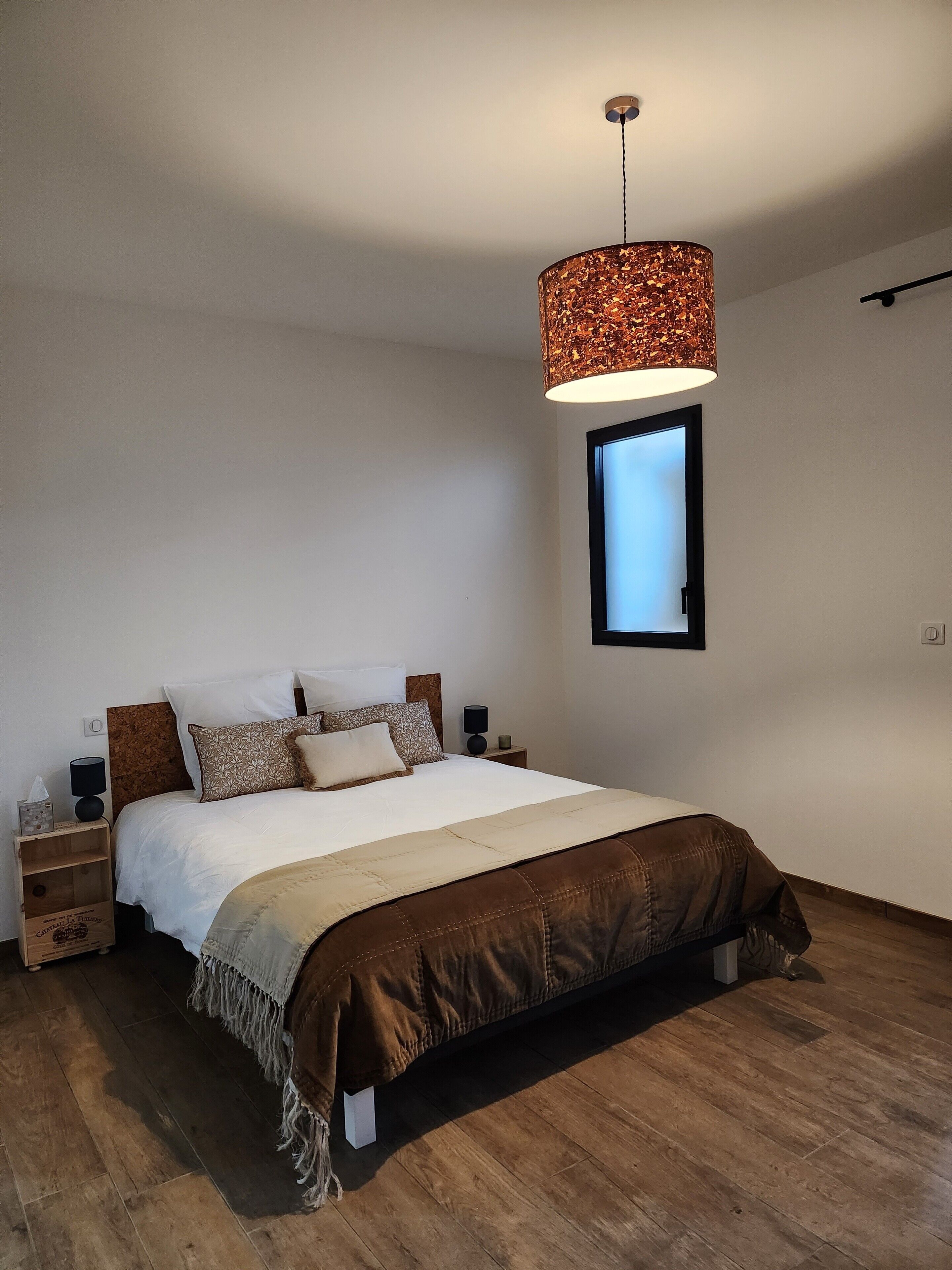 Un dormitorio con cama, mesitas de noche, lámpara colgante, ventana y suelo de madera.