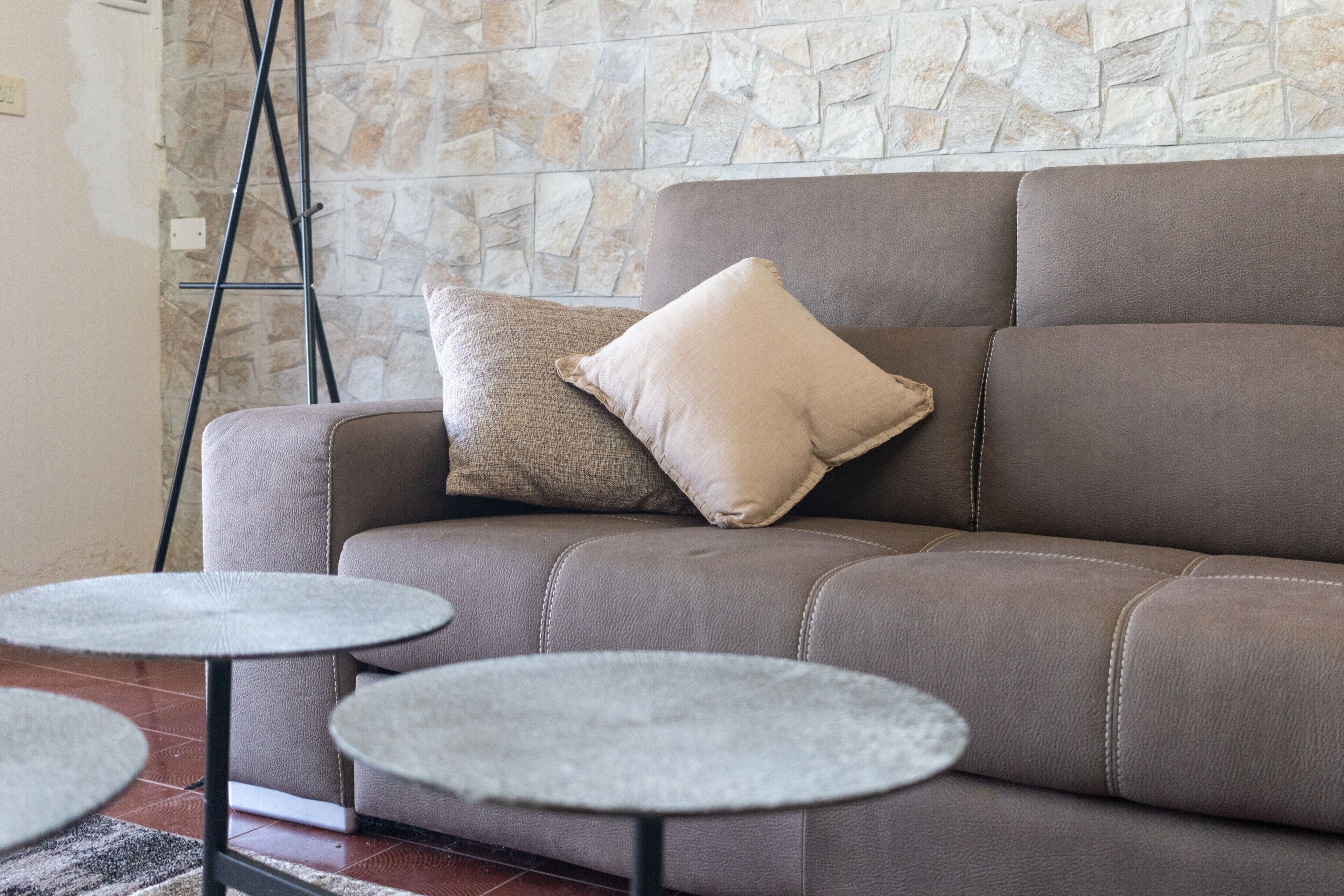 A brown leather sofa with a beige pillow against a stone wall, accompanied by two small round tables.