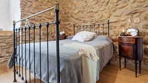 Ein Zimmer mit Steinwand, einem Bett mit grauer Decke, einem Nachttisch mit einer Uhr und einem Beistelltisch mit einer Lampe.