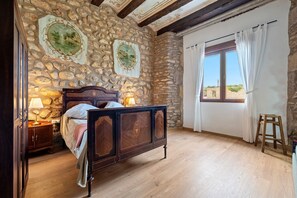 A bedroom with a stone wall, wooden bed, nightstand, lamp, and a window with curtains.