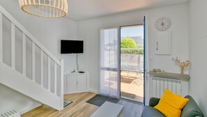 A modern living room with a staircase, a television, a sliding glass door to an outdoor area, and a clock on the wall. - Holiday Home 'Petite Maison' with Private Terrace and Wi-Fi (Le Croisic)