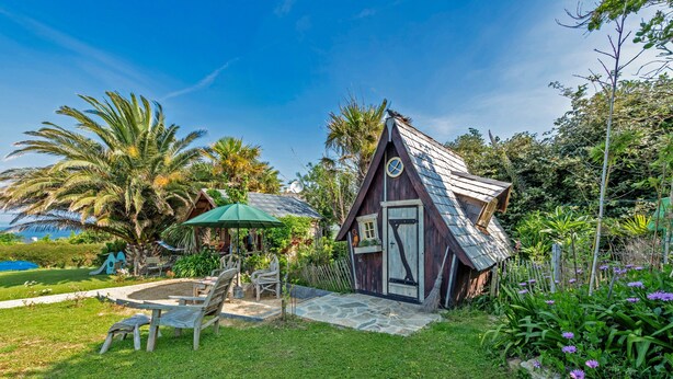 A small wooden cabin with a sloped roof, surrounded by a garden with palm trees and purple flowers. - Chalet 'With Garden and Sea View 100m' with Sea View, Private Terrace and Garden (Plougastel-Daoulas)