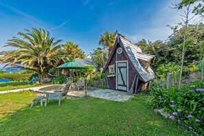 Une petite cabane en bois, dotée d’un toit en pente, entourée d’un jardin planté de palmiers et de fleurs violettes.