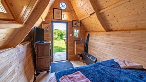A wooden cabin with a sloped ceiling, a bed with a blue blanket, a small desk with a television, and a bathroom with a sink and shower. - Chalet 'With Garden and Sea View 100m' with Sea View, Private Terrace and Garden (Plougastel-Daoulas)