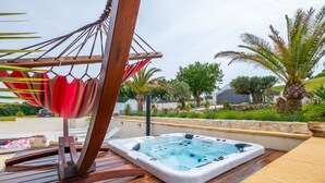 Un jacuzzi en una terraza de madera con una hamaca y vistas a la vegetación y colinas.