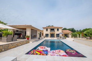 Une piscine avec des chaises longues et un mur en pierre, un patio couvert et une maison à deux étages avec un toit en tuiles.
