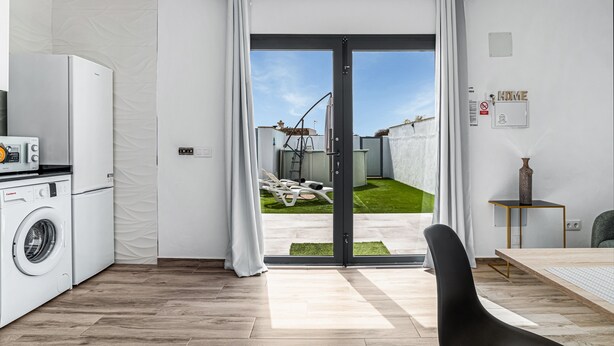 Een moderne keuken met een wasmachine, een eethoek met een tafel en stoelen, en een schuifdeur naar een terras met gazon en een ladder.