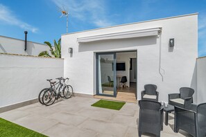 Un patio moderno con una pared blanca, un cerramiento de vidrio corredero que da a un interior, una mesa pequeña con sillas y bicicletas apoyadas contra la pared.