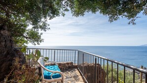 Una escalera de piedra conduce a un balcón con una silla de mimbre y cojines, con vistas a la costa.
