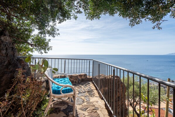 A stone staircase leading to a balcony with a wicker chair and cushions, overlooking a coastal view.