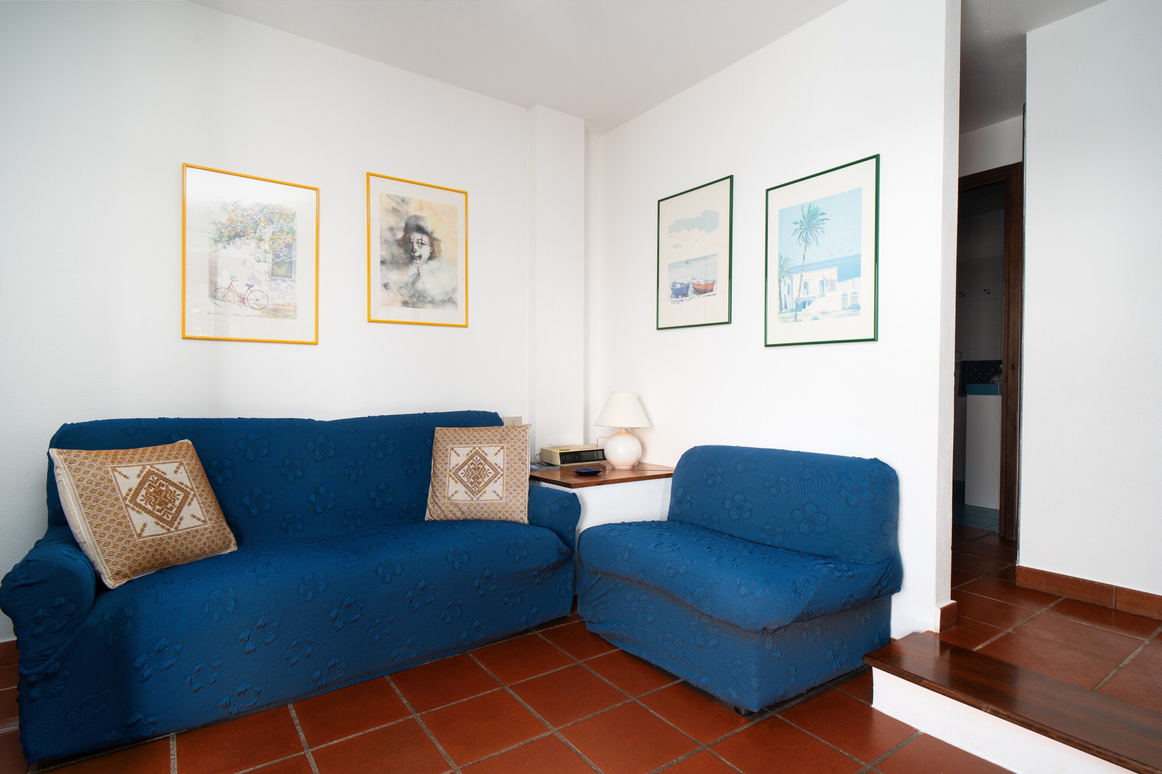 Una sala de estar con un sofá azul, una mesita con una lámpara y cuadros en la pared.