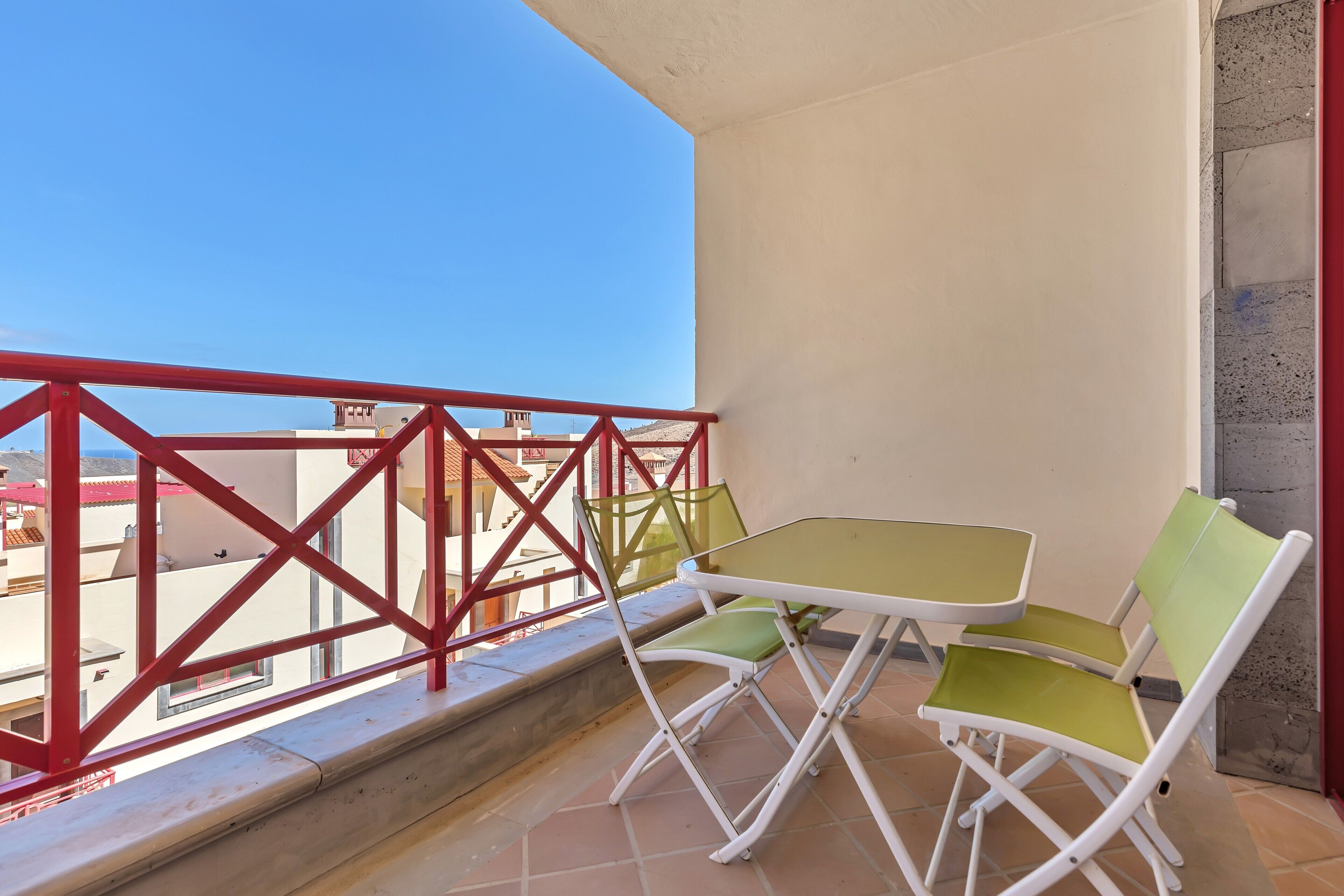 Een balkon met een rode reling, twee groene en witte stoelen en een tafeltje.