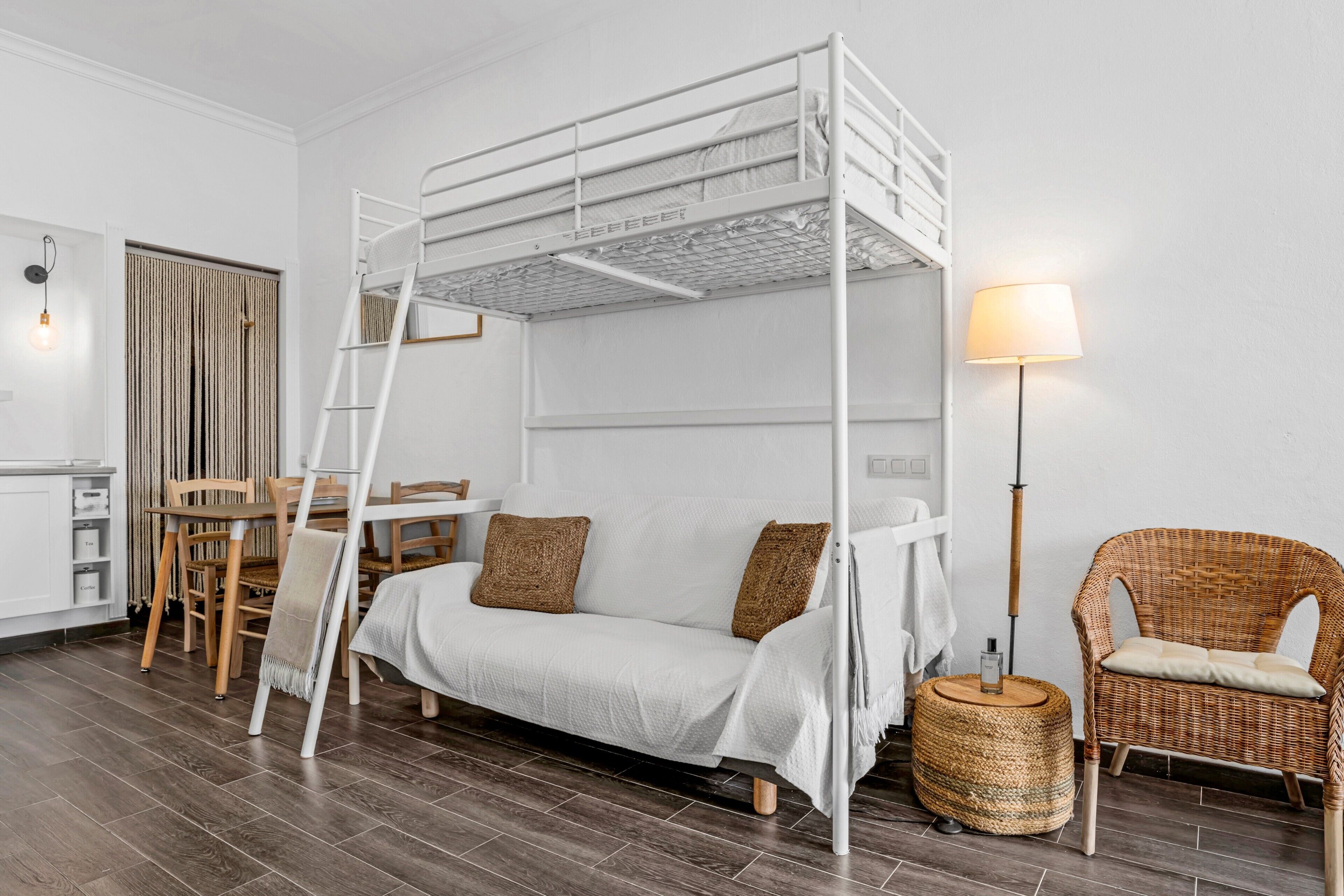 Un lit superposé avec une échelle, un canapé blanc avec des coussins, une chaise en osier, une petite table ronde et une lampe.