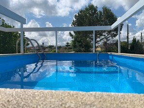 A clear blue swimming pool with a white deck and metal ladder, surrounded by trees and a clear sky.