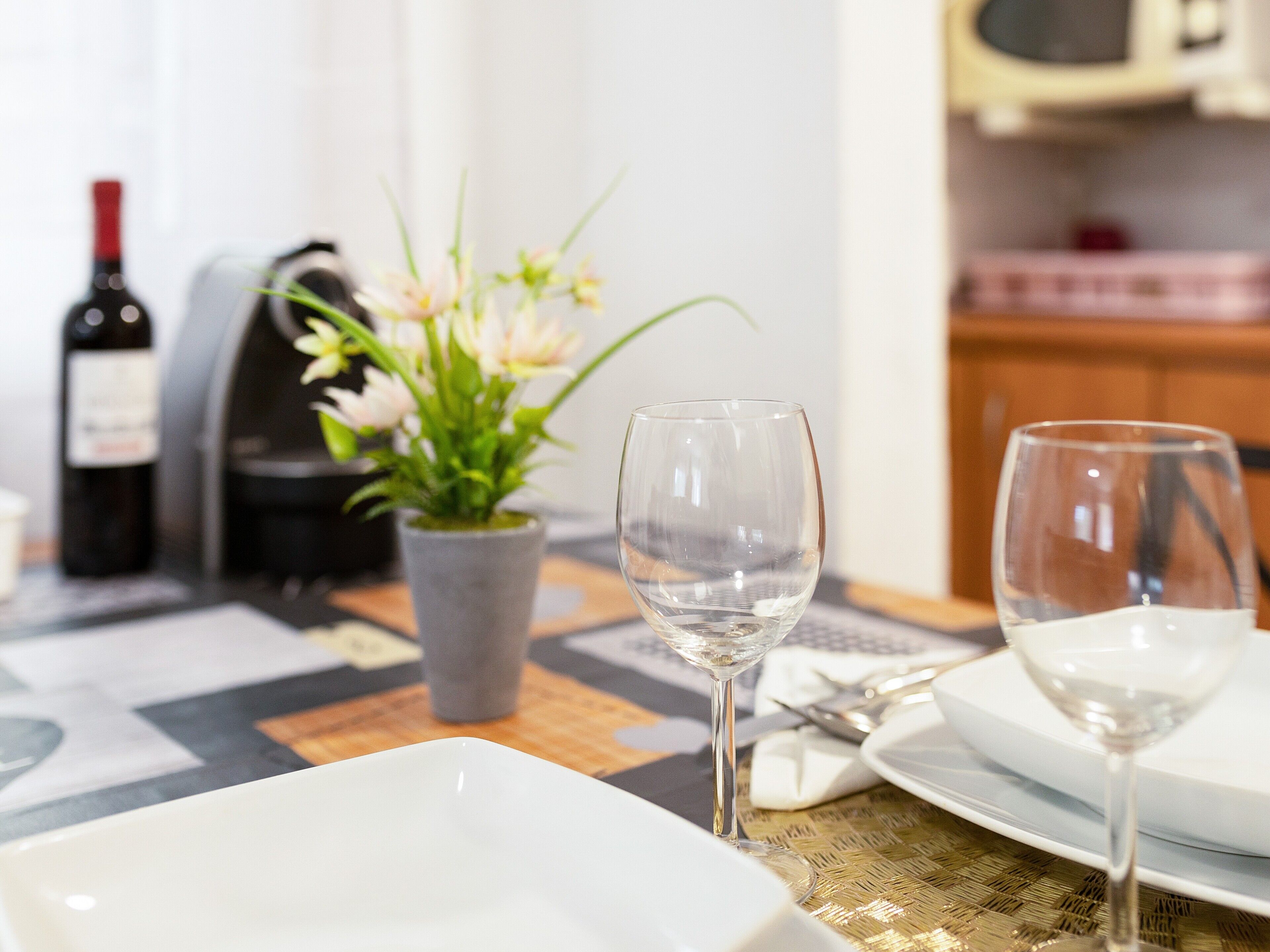 Una mesa de comedor con dos copas de vino, una botella, una planta en maceta y un microondas al fondo.