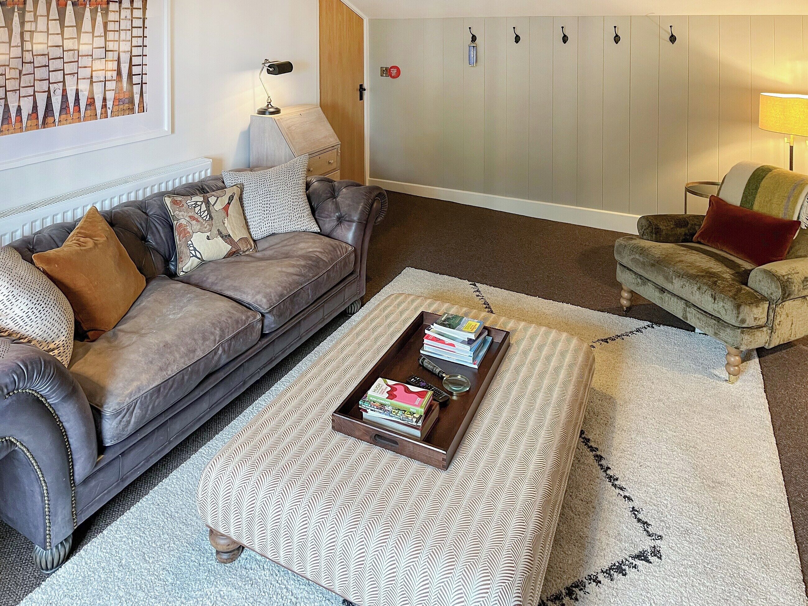 A living room with a grey sofa, a patterned ottoman, a wooden coffee table with books, a lamp, and a chair with a red cushion.