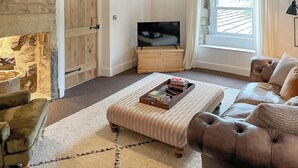 A cozy living room with a stone wall, a wooden door, a television, a sofa, and a patterned ottoman. - The Hermit Inn Hideout (Burley Woodhead, near Ilkley)