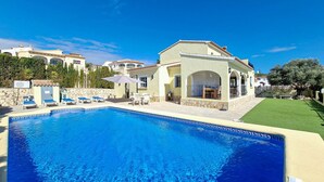 Vue sur une villa de style méditerranéen avec piscine, espace salon extérieur et pelouse bien entretenue, le tout vu depuis le bord de la piscine.