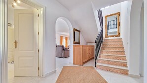 Un hall d’entrée spacieux avec une porte blanche, un meuble en bois, un tapis à motifs et un escalier doté de rampes décoratives.