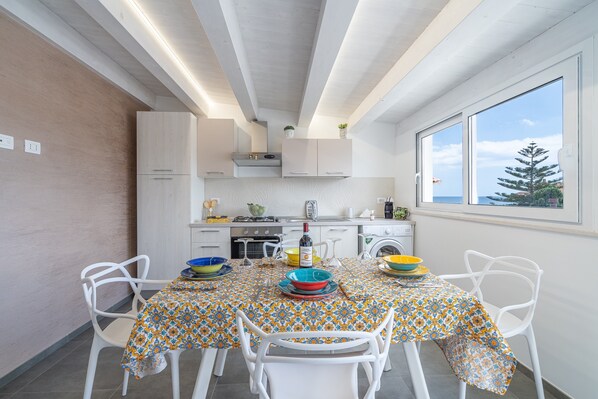 A modern kitchen with white cabinetry, a dining table with a patterned tablecloth, and a view of the sea through a large window.